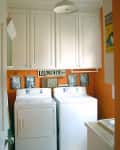 Laundry room with orange walls, white cabinets, washer and dryer, vintage laundry sign, and framed photos.