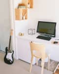 Home office with a wooden desk, computer, chair, wall shelves, and a black electric guitar leaning against the wall.