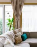 Cozy living room with a gray sectional sofa, beige curtains, green and beige cushions, and potted plants by large windows.