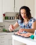 Woman decorating pink donuts in a white kitchen with floral blouse.