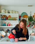 Woman pouring tea from a red kettle into striped mugs in a colorful kitchen with open shelves and vintage decor.