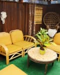 Bohemian sunroom with yellow wicker furniture, green plants, macramé decor, and a round mosaic table on a green rug.