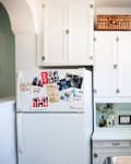 White kitchen with cabinets, fridge covered in photos and cards, potted plants on windowsill, and utensils hanging on yellow tiles.