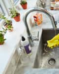 Person wearing yellow gloves cleaning a kitchen sink with a sponge, surrounded by potted plants and cleaning supplies.