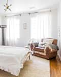 Bright bedroom with a white bed, green pillows, leather armchair, and wall art, featuring a plant and large windows.