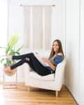 Woman sitting on a white armchair reading a magazine, with a plant and macrame wall hanging in a bright room.