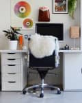 Home office with a white desk, black chair with fur throw, computer monitor, potted plant, and colorful wall art.