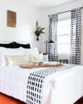 Bedroom with white bed, patterned pillows, black and white curtains, wall art, and a side table with decor.
