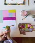 Person holding a paint bottle at a desk with colorful paper, paintbrushes, and a laptop.