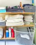 Laundry room shelves with folded towels, cleaning supplies, a wire basket, and decorative items.