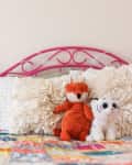Pink metal bed with ruffled pillows, orange fox and white tiger plush toys, colorful patchwork quilt, and books on a side table.