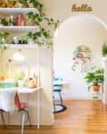 Cozy bedroom with red bed frame, plants, white desk, and archway leading to a room with a blue chair and round shelf.