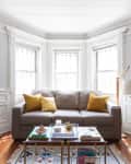 Bright living room with a gray sofa, yellow pillows, coffee table, and large windows. Shelves and plants add decor.