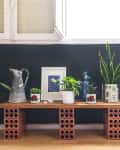 Indoor plant display on a wooden bench with brick supports, featuring various potted plants, a metal pitcher, and framed art.