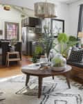 Living room with large plants, a wooden coffee table, gray sofa, and a modern chandelier near a kitchen area.