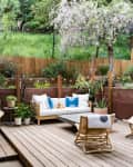 Outdoor patio with wooden deck, wicker furniture, hanging chair, potted plants, and a blooming tree in the background.