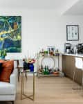 Modern living room with a white sofa, colorful cushions, bar cart, and kitchen area. Large painting and mirror on the wall.