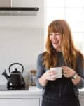 Woman in a kitchen holding a mug, with a black kettle on the stove and a potted plant by the window.