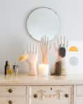 Vanity with decorative hand stands, jewelry, perfume bottles, a lit candle, and a round mirror on a white dresser.