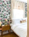 Bedroom with floral wallpaper, white bedding, wooden nightstand, and a gold lamp.