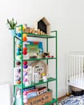 Nursery with a white crib, colorful geometric quilt, green shelf with toys and books, and multicolored bunting.