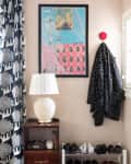 Framed artwork above a wooden side table with a lamp, patterned curtain, coat on hook, and shoe rack with various shoes.