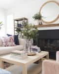 Living room with white sofa, pink and blue pillows, wooden coffee table, books, plants, and a round mirror above the fireplace.