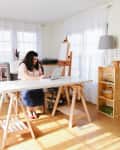 Woman working on a laptop at a white desk in a bright home office with bookshelves and an easel.