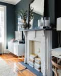 Dark-walled living room with ornate mirror, white fireplace, stacked books, and a TV on a white console.