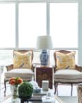 Living room with striped sofa, floral pillows, two armchairs, a blue and white lamp, and a stack of magazines by the window.