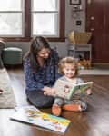 Mother and child reading on the floor in a cozy living room with a gray sofa and a basket of blankets.