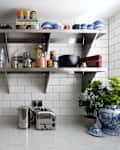 Kitchen counter with espresso machine, kettle, toaster, and open shelves holding glassware, jars, and blue-striped mugs.