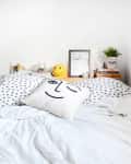 Cozy bedroom with eye-patterned bedding, smiley face pillow, cactus, and books on a wooden shelf.