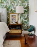 Bedroom with tropical leaf wallpaper, tufted headboard, colorful pillows, wooden nightstand, lamp, and vintage fan.
