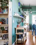 Bright kitchen with teal walls, wooden floor, hanging pots, and a person moving near white cabinets and appliances.