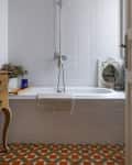 Bathroom with white tiled walls, a bathtub, overhead shower, patterned floor, and a wooden side table with decor.