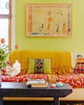 Cozy living room with a cream sofa, colorful cushions, yellow throw, floral vase, and books on a floating shelf.