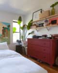 Cozy bedroom with white bedding, red dresser, plants, and wall art between two windows.