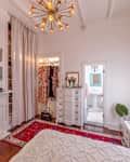 Bedroom with a red rug, white dresser, floral decor, and a modern chandelier, leading to a bathroom with a pedestal sink.