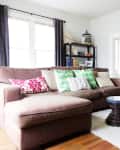 Brown sectional sofa with colorful pillows, black armchair, wooden tables, and bookshelf in a bright living room.