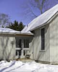 Snow-covered yard with a wooden chair on a porch outside a light-colored house.