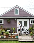 Small purple house with a wooden deck, potted plants, string lights, and a person with a bicycle and garden hose.