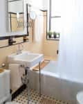 Small bathroom with beige tiles, white sink, and bathtub. A shower curtain, wall mirror, and potted plants on the window ledge.