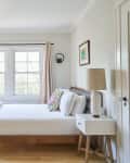 Bright bedroom with a wooden bed, white bedding, colorful pillows, bedside table, lamp, and large windows with beige curtains.