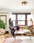 Three people in a bright living room with a brown sofa, large plant, abstract art, and a coffee table with a white bowl.