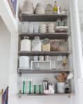 Open kitchen shelves with stacked plates, glass jars, mugs, wine glasses, and a cocktail shaker.