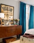 Mid-century modern bedroom with blue curtains, a wooden sideboard, eclectic pillows, and a vintage poster.