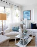 Bright living room with white sectional sofa, blue and beige pillows, glass coffee table, and large windows with sheer curtains.