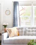 Gray tufted sofa with striped orange pillows, blue curtains, and a floor lamp in a bright living room.