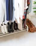Clothing rack with assorted garments, shoes, a brown leather bag, and a potted plant on a carpeted floor.